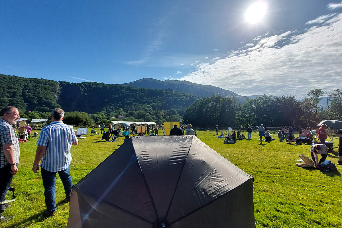 Artists setting up their kit in a field in the bright morning sun. The landscape in the distance shows a mountainous and lush hill side covered in trees with a distinctive quarry cut deep into the mountain side.